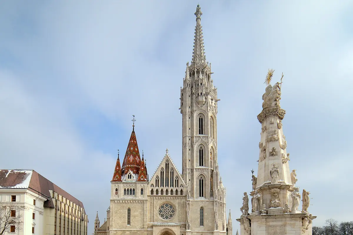 Matthias Church on a snowy square with its tall spire, tiled gables and the white Holy Trinity Column to the right